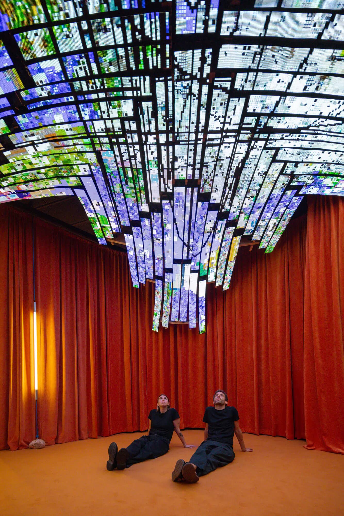 Dos personas sentadas en el suelo miran hacia arriba el domo LED de Simbiopolis, en el Palau Robert, con cortinas naranjas alrededor.